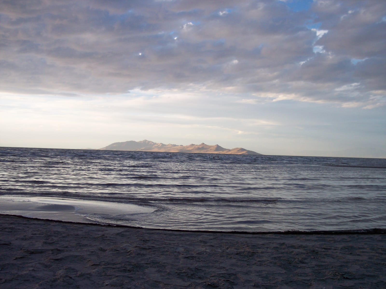 Small waves on Great Salt Lake at Sunset Beach under a cloudy sky, with Antelope Island on the horizon