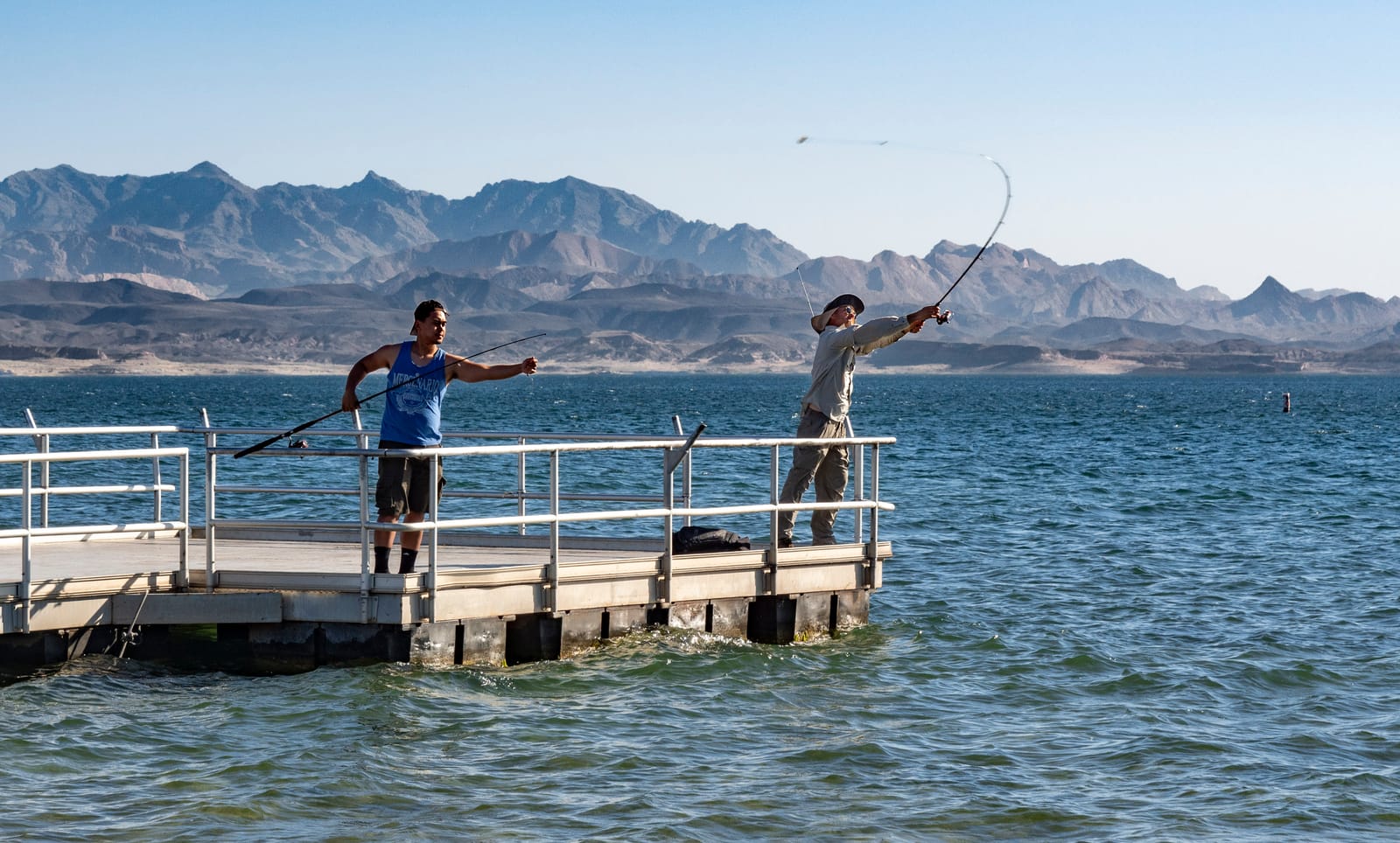 Two people casting from the Hemenway Harbor fishing pier on Lake Mead, with jagged desert mountains across the water