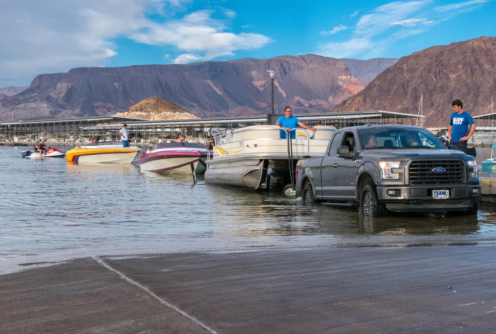 A pickup truck backing a boat trailer down the Hemenway Harbor launch ramp on Lake Mead, boats waiting in the water and desert mountains behind